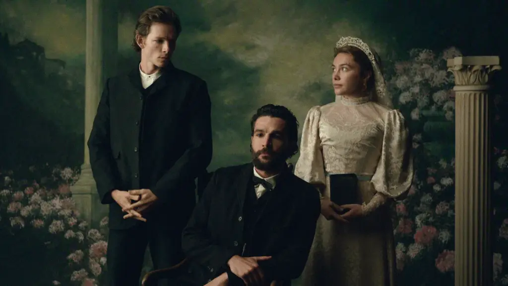 "Three people in Victorian-era clothing pose in a formal portrait setting with floral and classical column background; one man seated, one man standing, and a woman in a white dress holding a book."